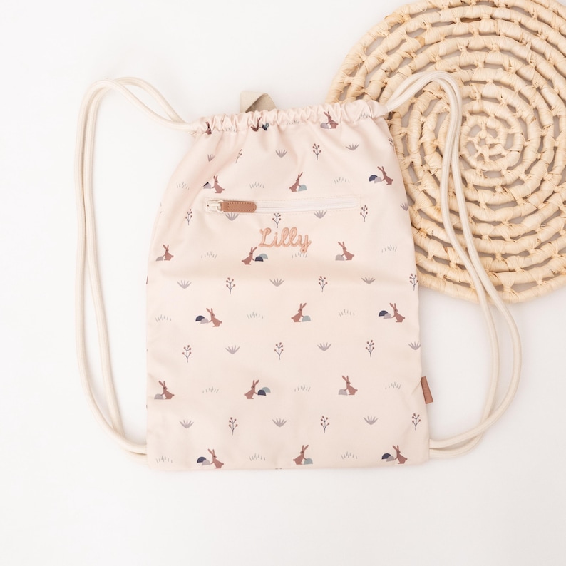 May include: A beige drawstring backpack with a repeating pattern of small rabbits, grass, and flowers. The backpack has a front zipper pocket and the name "Lilly" embroidered on it. The bag has white drawstrings and a woven circular mat is in the background.