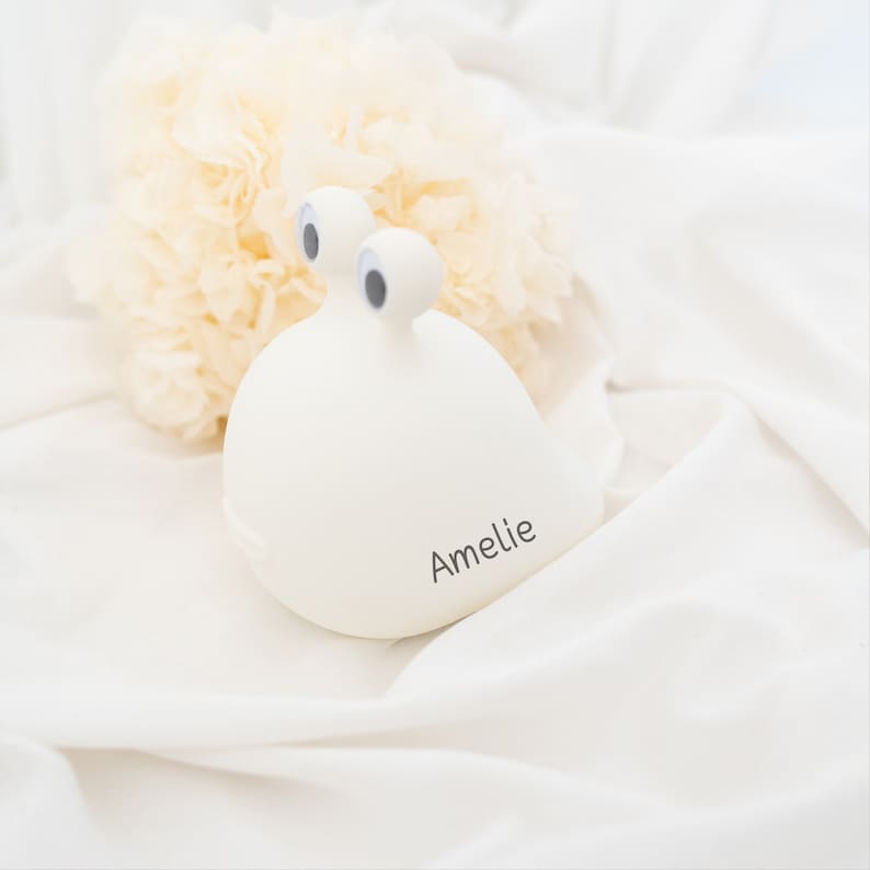 May include: A smooth, off-white, rounded snail-shaped object with two protruding stalks topped with black and white googly eyes. The side of the object features the name Amelie printed in a simple, dark, sans-serif font.