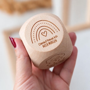 Puede incluir: Un cubo de madera de color claro con un diseño grabado con láser de un arcoíris con un pequeño corazón debajo. Debajo del arcoíris, está grabado el texto en alemán "Darauf freue ich MICH MORGEN". Un segundo lado muestra una nube con gotas.