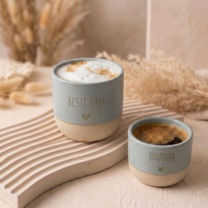 Pode incluir: Duas canecas de cer&acirc;mica com uma sec&ccedil;&atilde;o superior azul-clara salpicada e uma base bege mate. A caneca maior apresenta o texto 'BESTE OMA' com um pequeno &iacute;cone de cora&ccedil;&atilde;o, enquanto a menor tem o nome 'JOHANNA' e um &iacute;cone de cora&ccedil;&atilde;o.