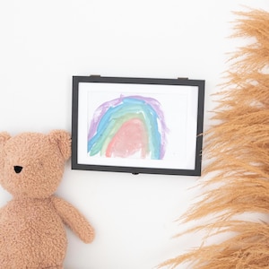 May include: A framed watercolour painting of a rainbow in shades of pink, blue, green, and purple. The painting is displayed on a white wall next to a brown teddy bear.