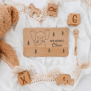 May include: A wooden tray with a black and white illustration of an elephant and the text "HIER KRUMELT Clara". The tray is surrounded by wooden blocks, a wooden spoon, and a stuffed animal.