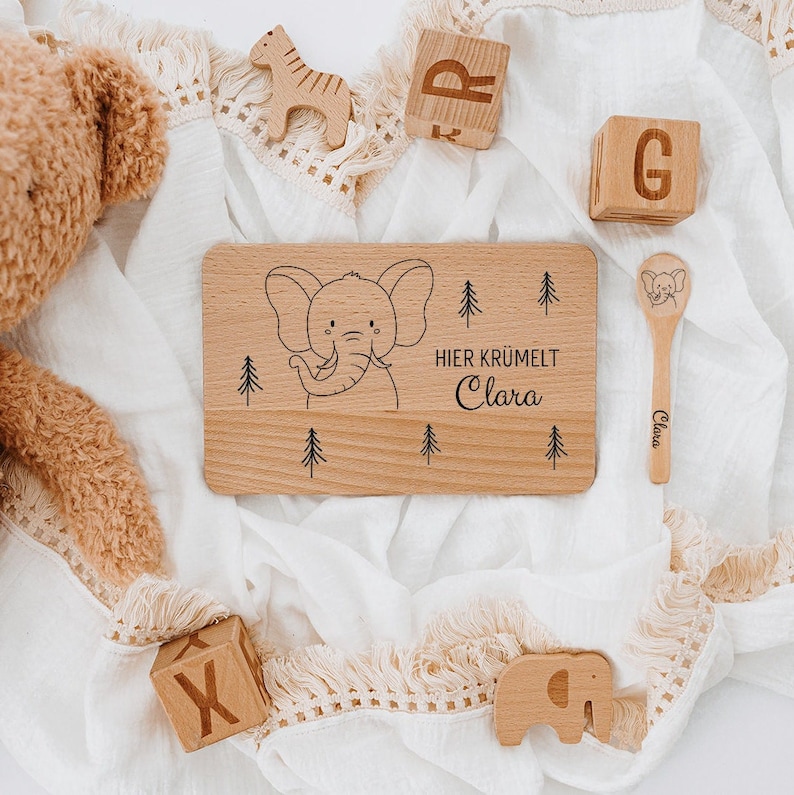May include: A wooden tray with a black and white illustration of an elephant and the text "HIER KRUMELT Clara". The tray is surrounded by wooden blocks, a wooden spoon, and a stuffed animal.