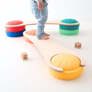 Può includere: Un set di travi di equilibrio in legno con assi in compensato chiaro collegate a basi circolari testurizzate di colore rosso, blu, verde e giallo. Due piccoli blocchi di legno con le lettere G e A sono posizionati sul pavimento.