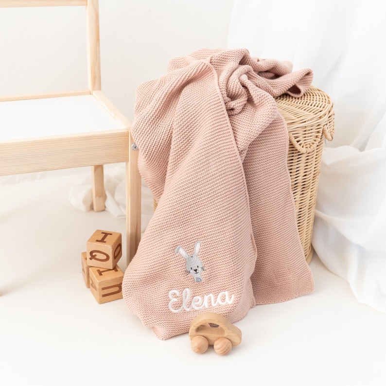 Puede incluir: Una manta de beb&eacute; de punto rosa claro con un conejito bordado y el nombre "Elena" en escritura blanca. La manta est&aacute; sobre una cesta de mimbre. Bloques de madera y un coche de madera en primer plano.