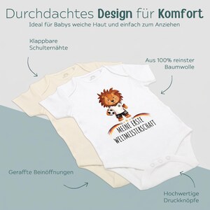Może przedstawiać: Dwa body dla niemowląt, jedno w kolorze kremowym i jedno białe, z napisem "MEINE ERSTE WELTMEISTERSCHAFT" i ilustracją lwa. Posiadają zapięcia na zatrzaski, marszczone otwory na nogi i składane szwy na ramionach.