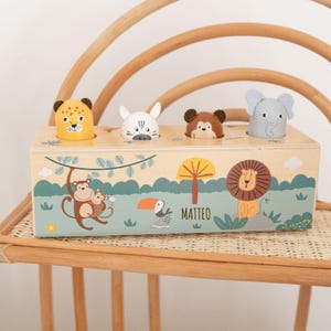 Könnte beinhalten: Holz-Pop-up-Spielzeug mit Dschungel-Thema, mit einem Leoparden, Kaninchen, Bären und Elefanten. Die Box hat eine Dschungel-Szene mit einem Affen, Tukan und Löwen. Der Name "MATTEO" ist aufgedruckt.