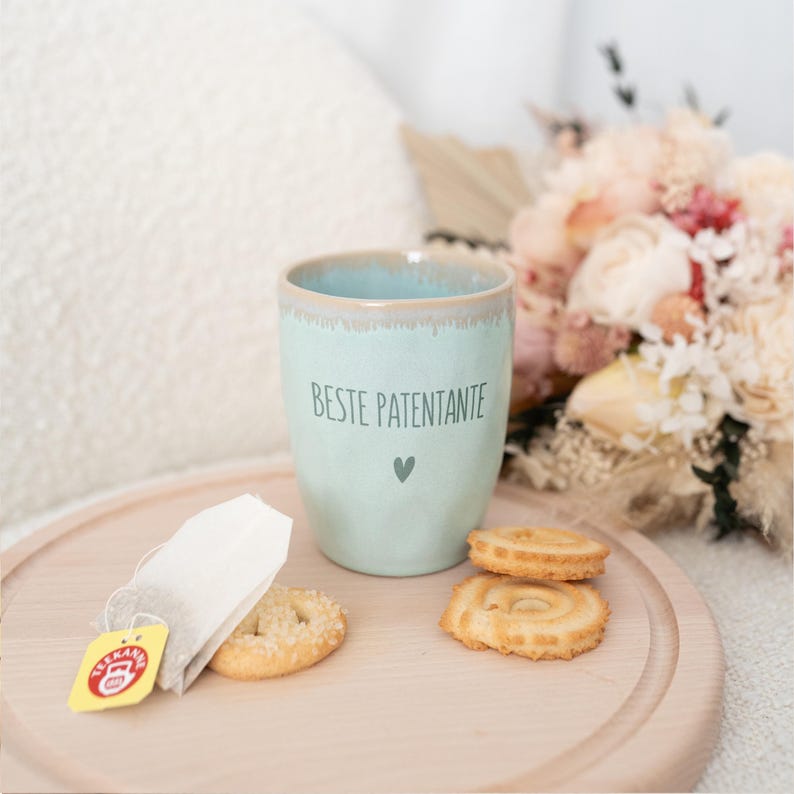 Puede incluir: Una taza de cer&aacute;mica azul claro con el texto "BESTE PATENTANTE" y un peque&ntilde;o coraz&oacute;n. La taza est&aacute; sobre una bandeja de madera con galletas y una bolsita de t&eacute;. Un ramo de flores est&aacute; en el fondo.