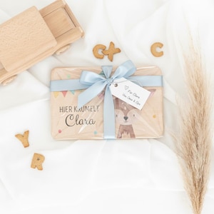 May include: A rectangular wooden board featuring a printed fox illustration, the text "HIER KRUMELT Clara," and a light blue ribbon tied in a bow. A white gift tag attached to the ribbon reads "F&uuml;r Clara Von Oma & Opa." Wooden toy truck and letter cookies.