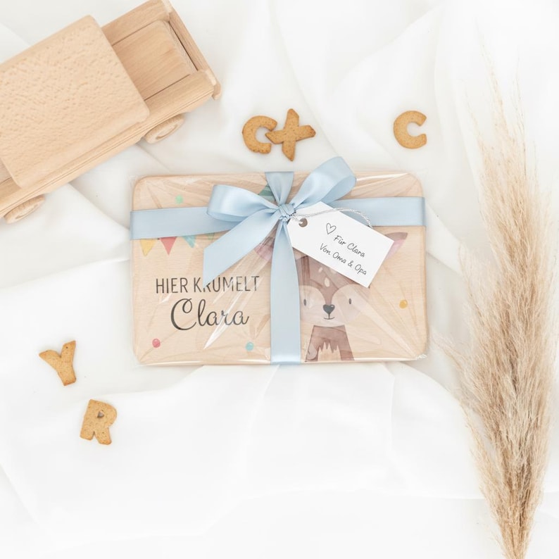 May include: A rectangular wooden board featuring a printed fox illustration, the text "HIER KRUMELT Clara," and a light blue ribbon tied in a bow. A white gift tag attached to the ribbon reads "F&uuml;r Clara Von Oma & Opa." Wooden toy truck and letter cookies.