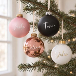 May include: Four spherical Christmas ornaments hanging from ribbons. One is matte pink with the name Hele, one is matte black with the name Toni, one is metallic rose gold with the name Celina, and one is glossy white with the name Julie in gold.