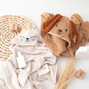 Könnte beinhalten: Zwei Kapuzenhandtücher, eines in Creme mit gesticktem Katzengesicht und dem Namen Mara, und eines in Braun mit gesticktem Hundegesicht, Schlappohren und dem Namen Pepe. Die Handtücher sind mit einer weißen Flasche und Holzbuchstaben abgebildet.