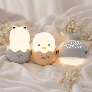 May include: Three illuminated night lights: a gray frog in an egg shell labeled THEO, a beige chick in an egg shell labeled EMILIA, and a gray whale with a star-patterned top that projects light. Each lamp emits a soft, warm glow.