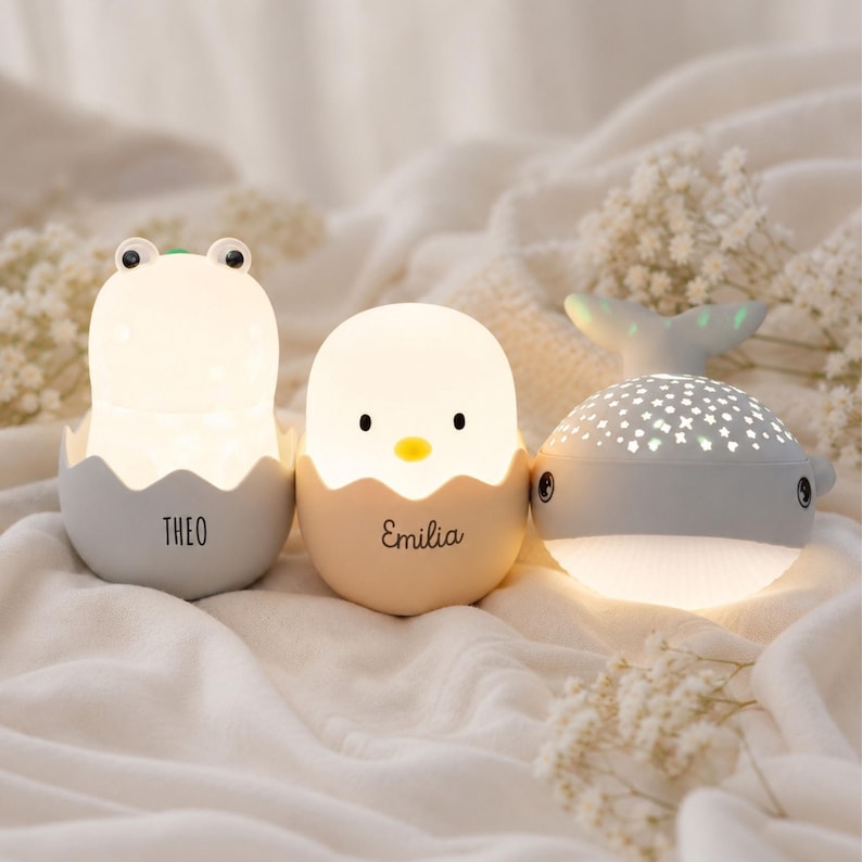 May include: Three illuminated night lights: a gray frog in an egg shell labeled THEO, a beige chick in an egg shell labeled EMILIA, and a gray whale with a star-patterned top that projects light. Each lamp emits a soft, warm glow.