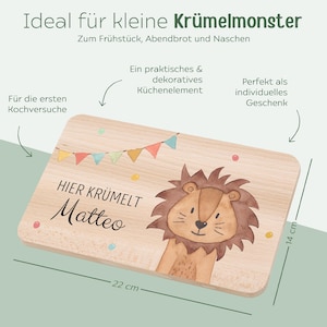 May include: A rectangular wooden cutting board measuring 22 cm by 14 cm. The board features a watercolor illustration of a lion, a colorful bunting banner, and the printed text "HIER KR&Uuml;MELT Matteo". The wood has a light, natural grain finish.