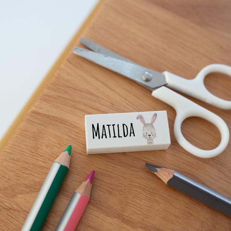 May include: A white rectangular eraser featuring the name Matilda in black sans-serif font next to a small illustration of a grey rabbit. The eraser is positioned near a pair of white-handled metal scissors and two coloured pencils.