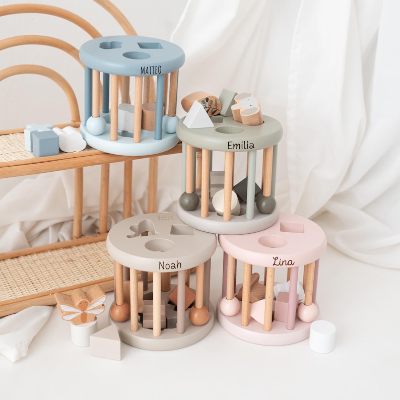 May include: Four wooden toy activity centers with different colored bases and personalized names: Matteo, Emilia, Noah, and Lina. Each center features a variety of shapes and colors for children to explore.