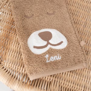 May include: A brown towel with a white embroidered dog face and the name "Leni" stitched in white.