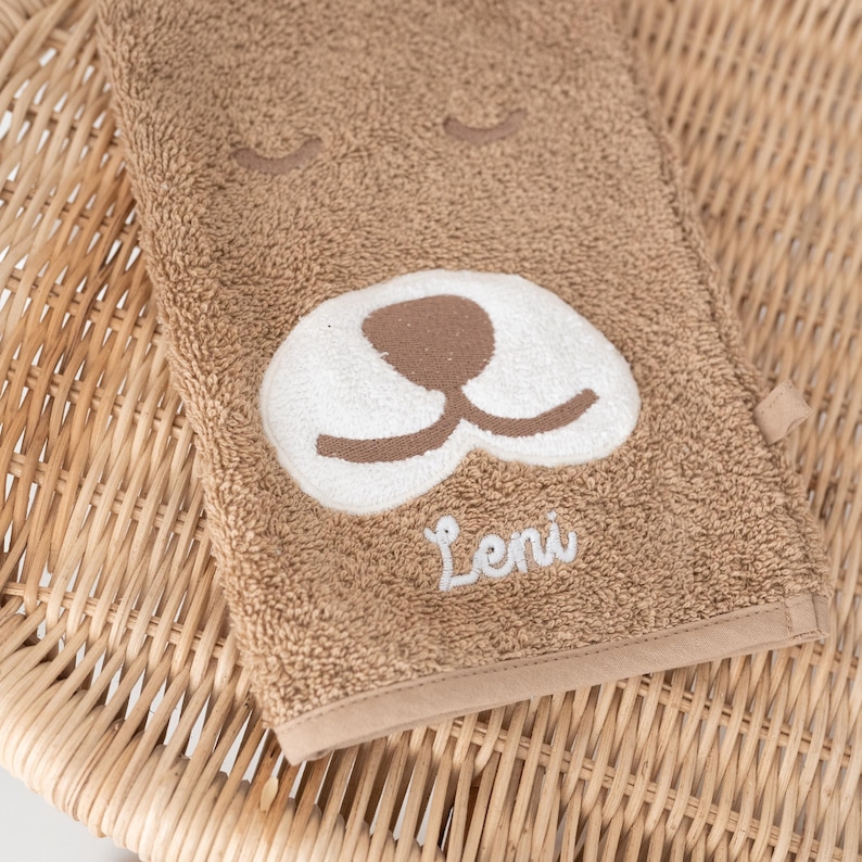 May include: A brown towel with a white embroidered dog face and the name "Leni" stitched in white.