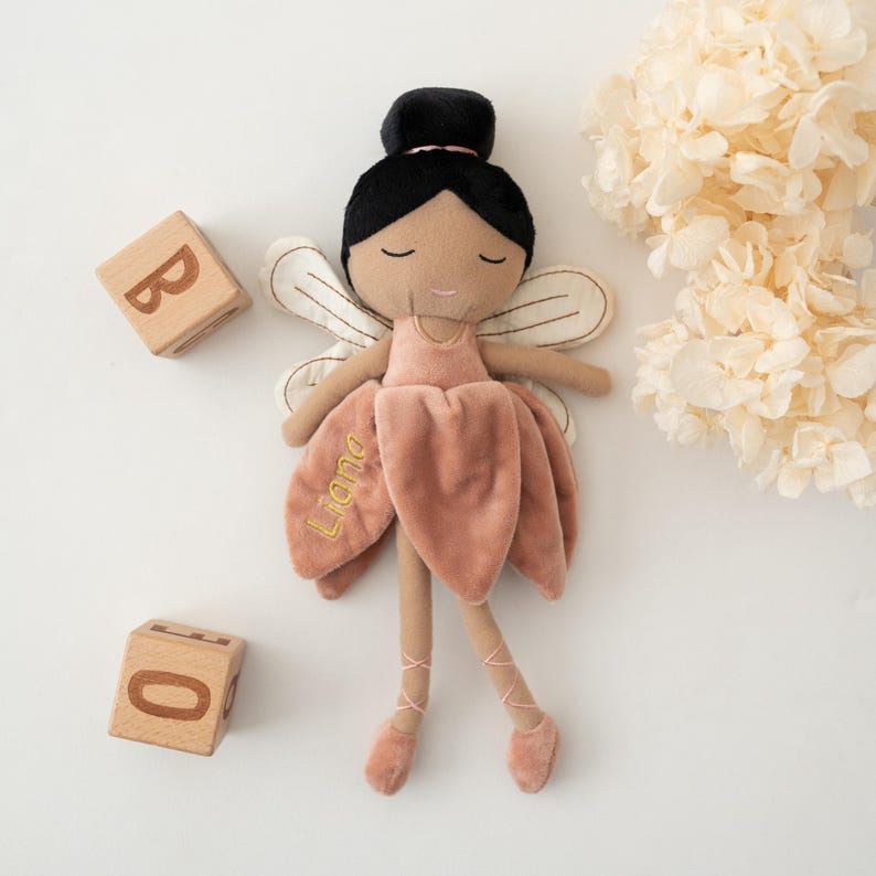 May include: A plush fairy doll with black hair, a pink dress, and embroidered details. The doll has a velvet skirt with the name "Liana" embroidered on it. Wooden blocks with the letters "B" and "O" are next to the doll.