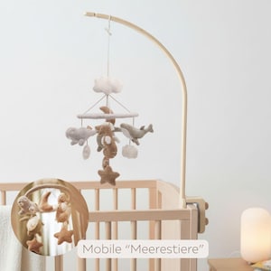 May include: A baby mobile with felted sea creatures, clouds, and stars in neutral tones hangs above a wooden crib. The mobile is suspended from a curved wooden arm. The text "Mobile 'Meerestiere'" is visible.