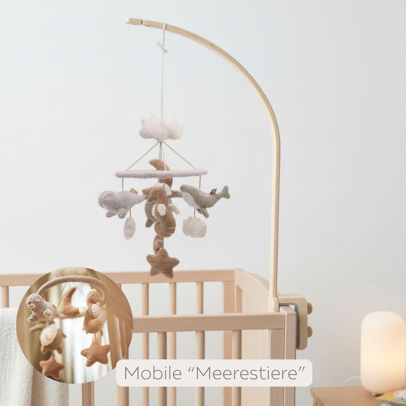 May include: A baby mobile with felted sea creatures, clouds, and stars in neutral tones hangs above a wooden crib. The mobile is suspended from a curved wooden arm. The text "Mobile 'Meerestiere'" is visible.