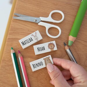 May include: Three rectangular white labels featuring black text and animal illustrations. One label reads Matilda with a rabbit, one reads Noah with a lion, and one reads Benno with a bear. The labels are arranged near white scissors and coloured pencils.