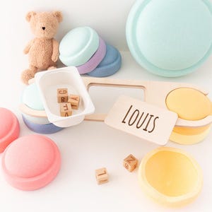 Può includere: Una collezione di giocattoli in legno dai colori pastello e un orsacchiotto. I giocattoli includono dischi impilabili, un vassoio con blocchi di lettere e un cartello di legno con la scritta "LOUIS". La tavolozza dei colori comprende rosa, giallo, blu e viola.