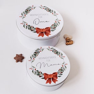 May include: Two white, round cookie tins with lids, decorated with a wreath design and a red bow. The top tin reads "WEIHNACHTSKEKSE F&Uuml;R Oma" and the bottom tin reads "WEIHNACHTSKEKSE F&Uuml;R Mama". A star-shaped biscuit is visible.