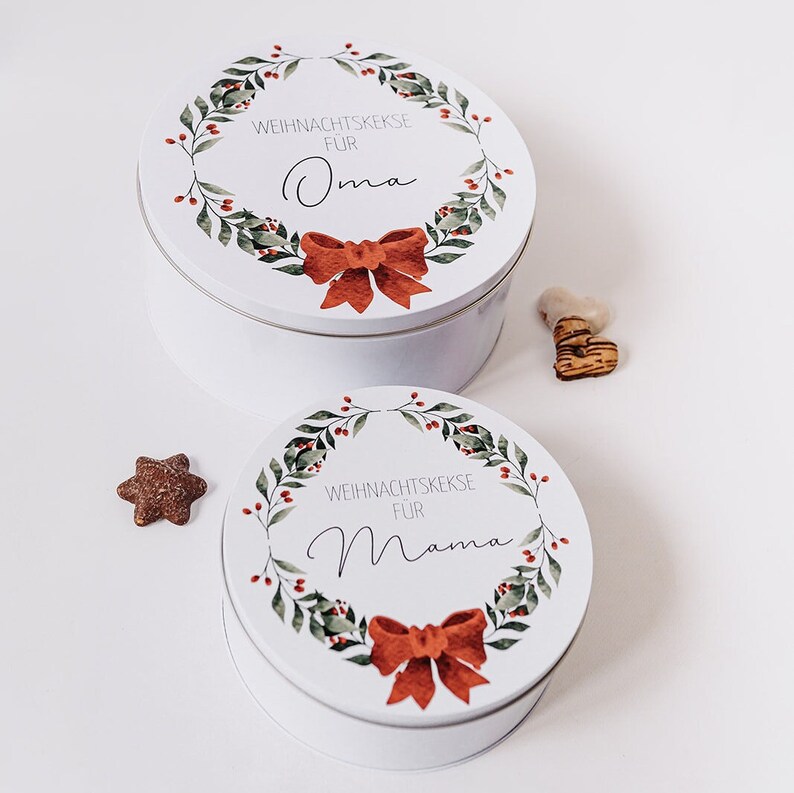 May include: Two white, round cookie tins with lids, decorated with a wreath design and a red bow. The top tin reads "WEIHNACHTSKEKSE F&Uuml;R Oma" and the bottom tin reads "WEIHNACHTSKEKSE F&Uuml;R Mama". A star-shaped biscuit is visible.