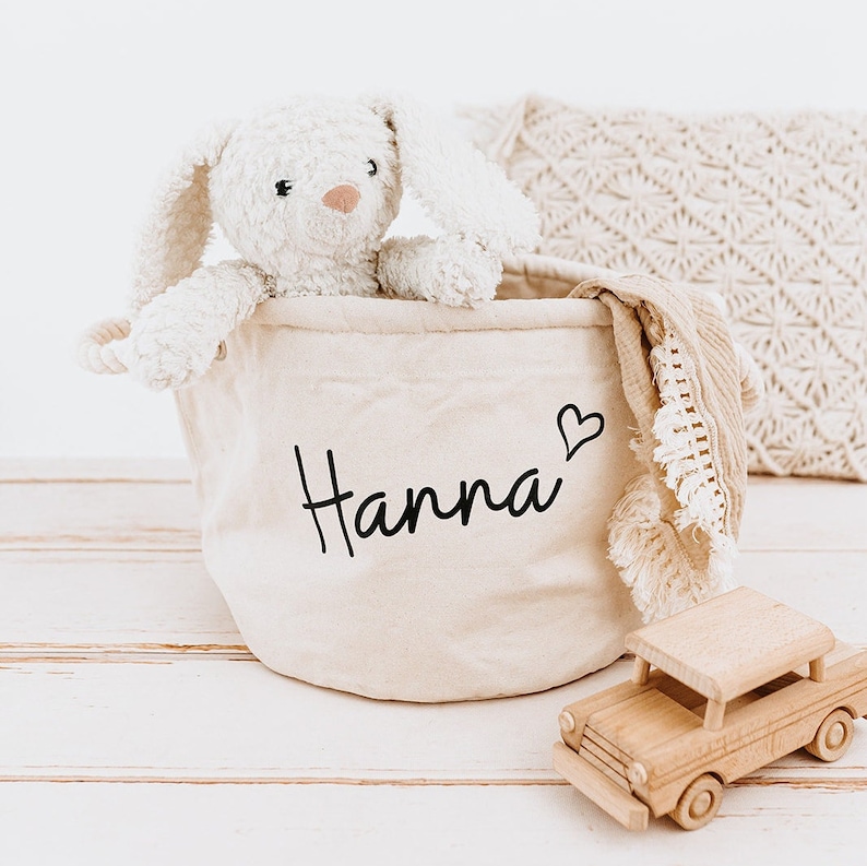 May include: A beige canvas storage basket with the name "Hanna" and a small heart embroidered on the front. The basket is filled with a white plush bunny, a beige blanket, and a wooden toy car.