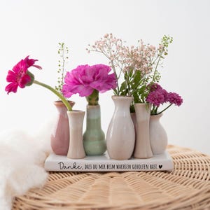 Può includere: Una collezione di piccoli vasi in ceramica in varie tonalità di rosa, beige e verde, che contengono vibranti fiori rosa e viola. I vasi sono disposti su un vassoio rettangolare bianco con testo in tedesco.