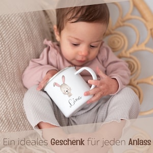 Puede incluir: Una taza de esmalte blanco con borde plateado que presenta una ilustraci&oacute;n en acuarela de un conejo marr&oacute;n y el nombre Lina impreso en letra cursiva negra. La taza tiene un asa curva y es sostenida por un ni&ntilde;o con una camisa rosa palo.