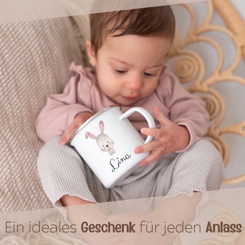 Puede incluir: Una taza de esmalte blanco con borde plateado que presenta una ilustraci&oacute;n en acuarela de un conejo marr&oacute;n y el nombre Lina impreso en letra cursiva negra. La taza tiene un asa curva y es sostenida por un ni&ntilde;o con una camisa rosa palo.