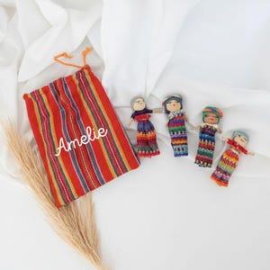 Peut inclure: Un sac à cordon rayé rouge, orange et jaune avec le nom "Amelie" en écriture blanche. Quatre petites poupées faites à la main avec des vêtements tissés colorés sont disposées à côté du sac. Les poupées ont des visages peints et portent des coiffes.