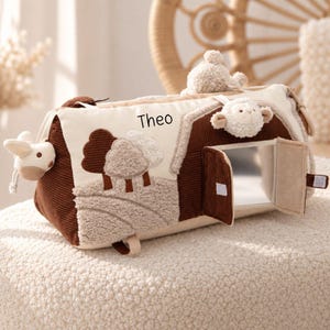 Peut inclure: Un jouet cylindrique en tissu représentant une grange avec des panneaux en velours côtelé marron et crème. Il comprend des accents en peluche en forme de mouton et de lapin, un miroir derrière une petite porte et le nom Theo en noir.