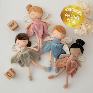 May include: Four plush fairy dolls with closed eyes, each with a different hair color and dress color: pink, blue, olive green, and peach. Each doll has embroidered wings and a name on the dress. Wooden blocks with letters and numbers are in the background.