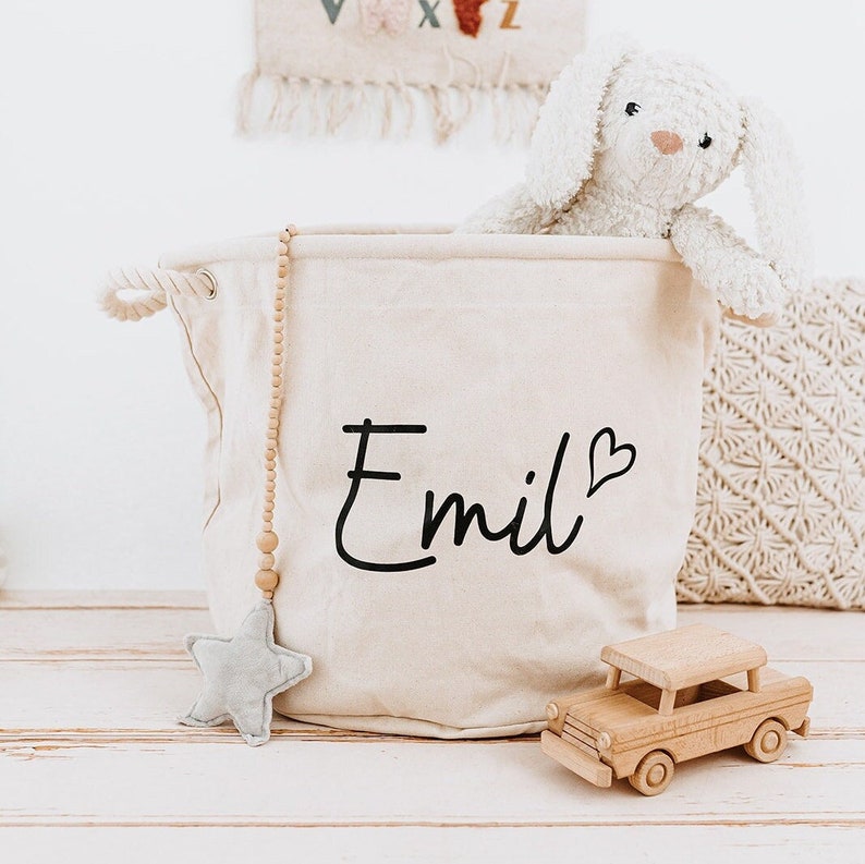 May include: A white canvas storage bin with the name "Emil" and a heart printed on the front. The bin has a wooden bead garland hanging from it and a gray star toy is sitting next to it. A wooden toy car and a white plush bunny are also in the image.