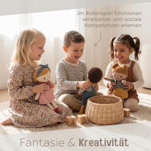 May include: Three children are smiling and playing with fabric dolls. The dolls have different skin tones and outfits. A woven basket and wooden blocks with letters are also in the image. Text at the top reads, "Im Rollenspiel Emotionen verarbeiten und soziale Kompetenzen erlernen." Text at the bottom reads, "Fantasie & Kreativit&auml;t."