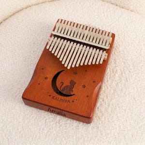 Könnte beinhalten: Eine hölzerne Kalimba mit siebzehn silberfarbenen Metallzungen. Das Instrument ist mit einer Lasergravur versehen, die eine Katze auf einer Mondsichel umgeben von Sternen zeigt. Unter dem Mond steht das Wort KALIMBA und der Name Amalia.