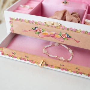 May include: A white wooden jewelry box with a pink interior and a floral design. The box has a drawer with a pink lining and a small gold knob. A white and pink beaded bracelet is in the drawer.