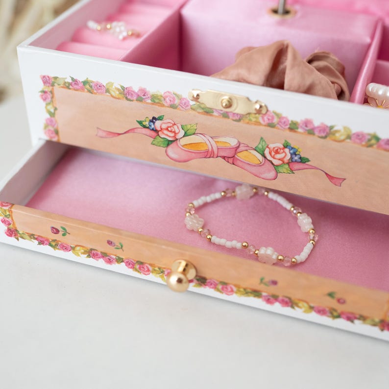 May include: A white wooden jewelry box with a pink interior and a floral design. The box has a drawer with a pink lining and a small gold knob. A white and pink beaded bracelet is in the drawer.
