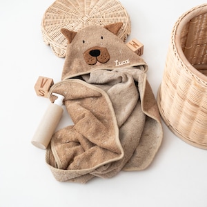 May include: A brown hooded baby towel with bear ears and a bear face applique. The towel is personalized with the name "Luca" embroidered on the hood. A beige bottle of soap and wooden blocks are next to the towel. A woven basket is in the background.