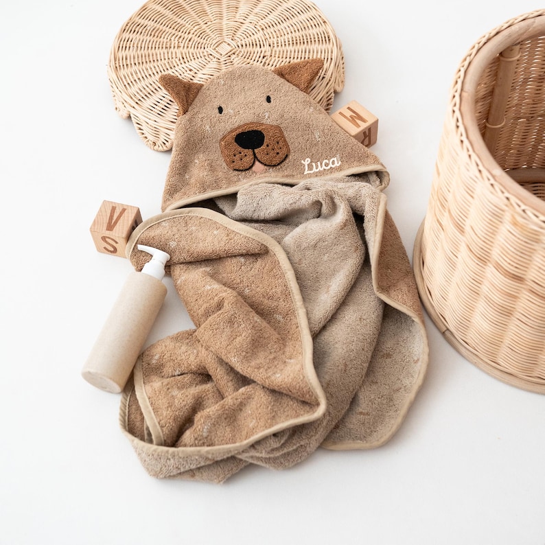 May include: A brown hooded baby towel with bear ears and a bear face applique. The towel is personalized with the name "Luca" embroidered on the hood. A beige bottle of soap and wooden blocks are next to the towel. A woven basket is in the background.