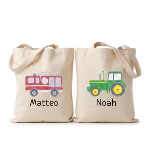 May include: Two beige canvas tote bags with handles. The left bag features a watercolor illustration of a red fire truck and the name Matteo in black text. The right bag features a watercolor illustration of a green tractor and the name Noah in black text.