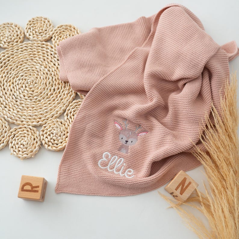 Puede incluir: Una manta de beb&eacute; de punto rosa empolvado con un dise&ntilde;o de ciervo bordado y el nombre "Ellie" en escritura blanca. La manta est&aacute; doblada, mostrando su textura. Bloques de madera con letras est&aacute;n cerca.