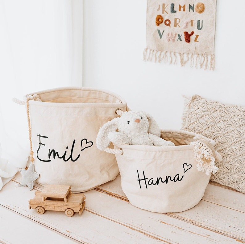 May include: Two beige canvas storage bins with rope handles. The larger bin has the name "Emil" written on it with a heart symbol. The smaller bin has the name "Hanna" written on it with a heart symbol. A white stuffed bunny is sitting in the smaller bin.