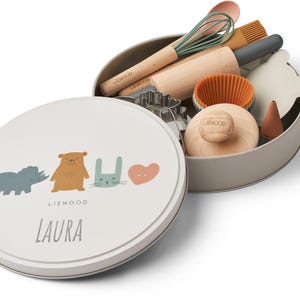 Könnte beinhalten: Eine hellgraue Metallbox mit Deckel, verziert mit Tierillustrationen und dem Namen "LAURA". Im Inneren befinden sich Backutensilien aus Holz und Silikon, darunter eine Teigrolle, ein Schneebesen und Ausstecher. Die Box ist geöffnet und zeigt den Inhalt.