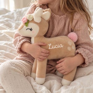 Peut inclure: Une peluche en forme de faon avec une fourrure beige, des détails blancs sur le visage et les sabots, et une queue en pompon rose. Le faon porte une couronne de fleurs en feutrine entre les oreilles et le prénom Lucia brodé en blanc.
