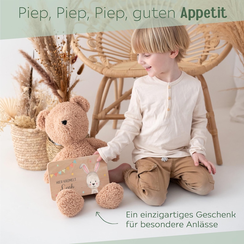 May include: A light brown plush teddy bear holding a rectangular wooden board with a printed illustration of a rabbit, colorful bunting flags, and the text "Hier kr&uuml;melt Leah". The bear has dark eyes and a soft, textured fabric finish.